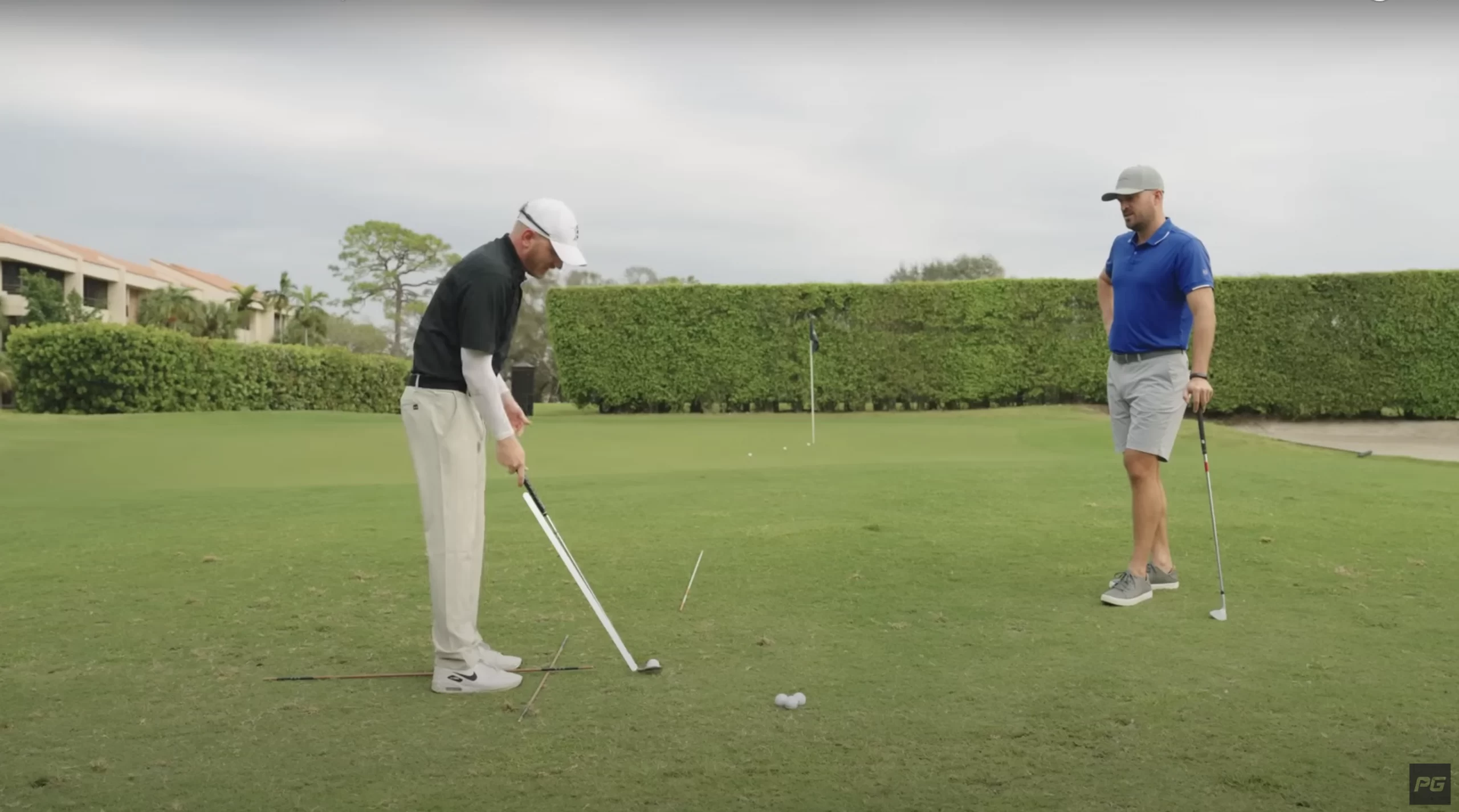 Two Performance Golf coaches on the green, one on the left is in the golf short game setup while the other is on the right, leaning on his golf club watching on.