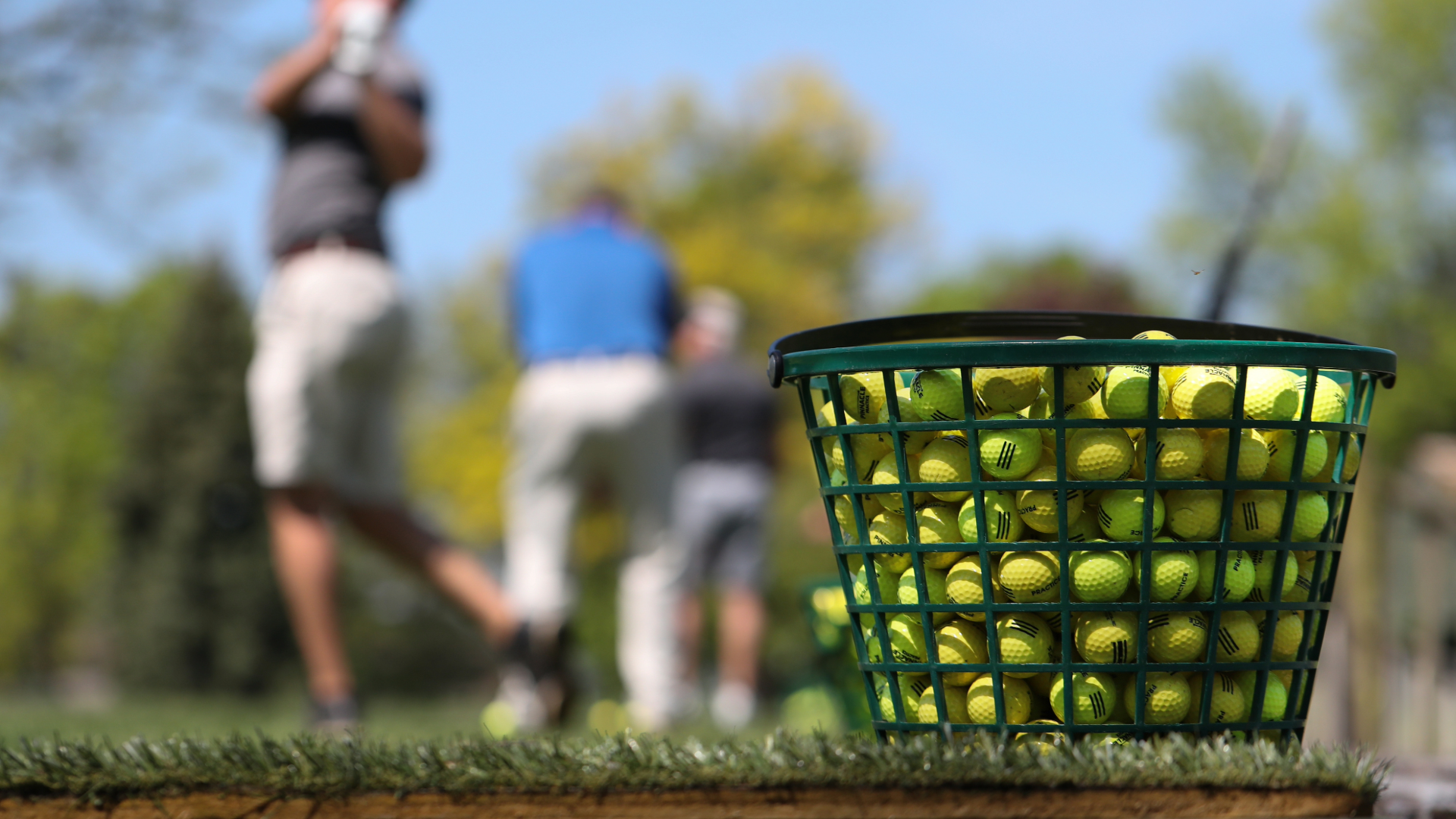 Close-up of a bucket full of yellow golf balls, with various golfers in the background practicing their golf swings.